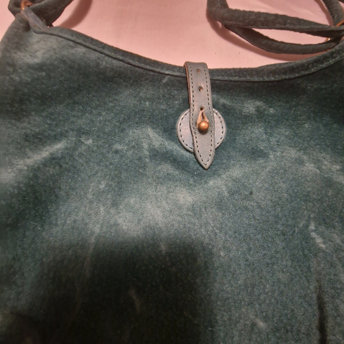 Close-up of the teal suede bag front showing the pointed leather tab fastened over a brass-tone stud