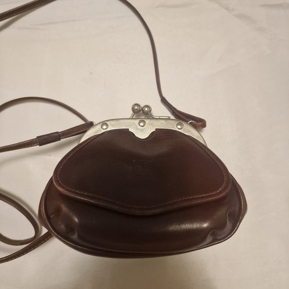 Dark brown leather kiss-lock crossbody bag lying flat with silver-tone frame, ball clasp and long thin strap