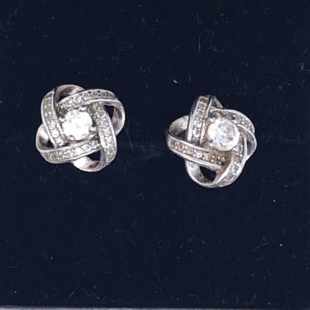 Straight view of two silver-tone interlaced knot earrings with pave bands and a clear centre stone