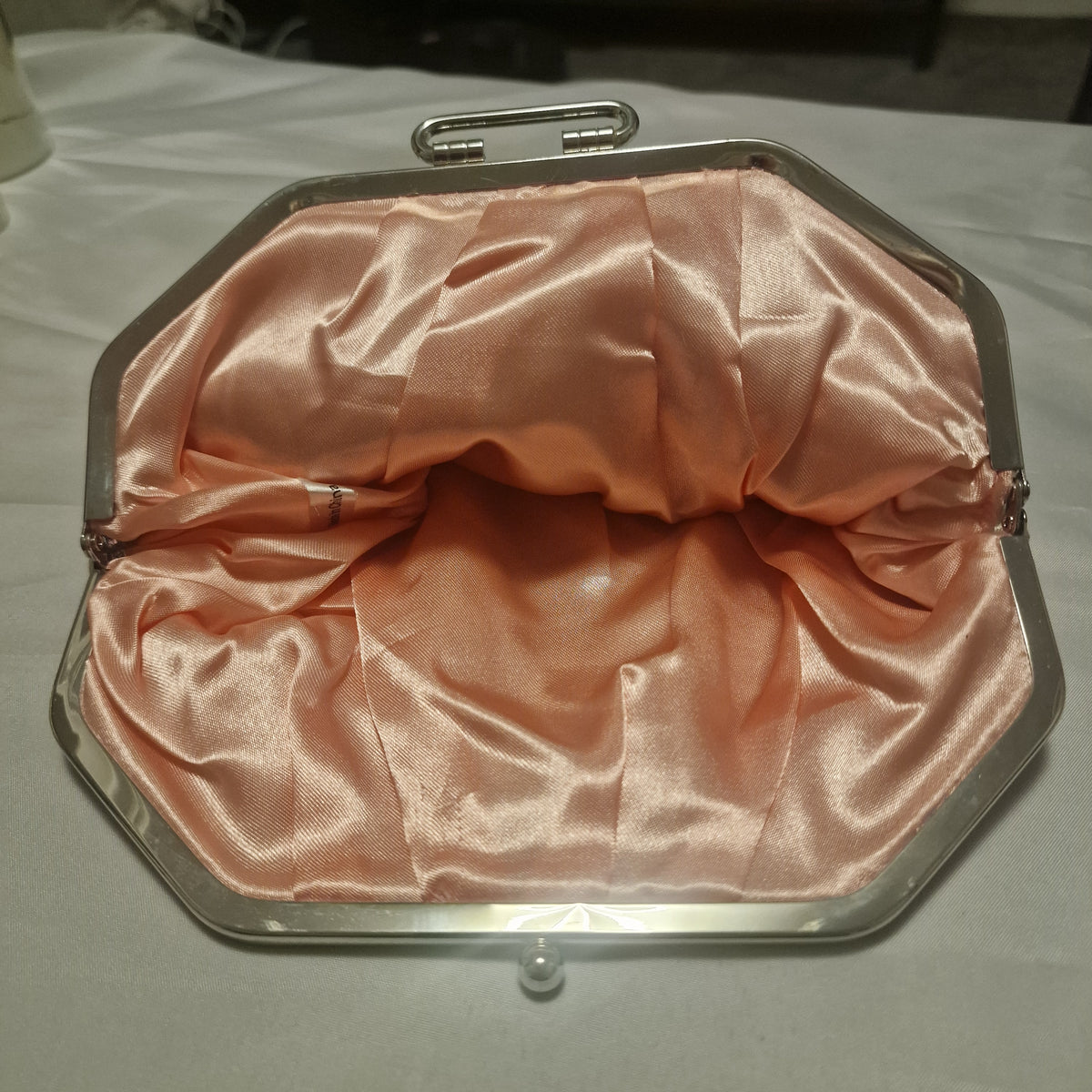 Open clutch purse showing its shiny peach-pink lining, metal frame, and a partly obscured interior label.