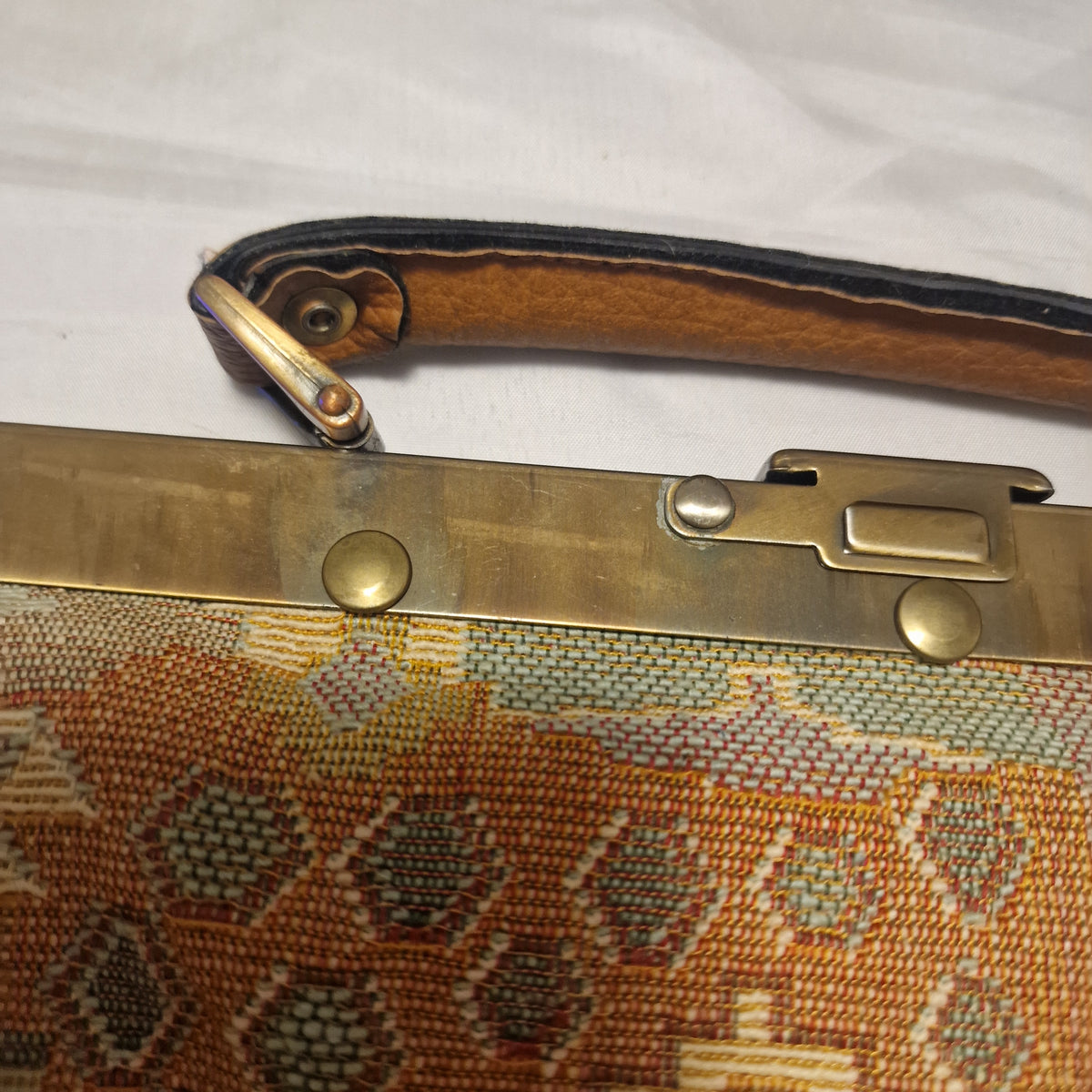 Brass-tone slide clasp and rivets on the frame next to the tan handle and tapestry fabric
